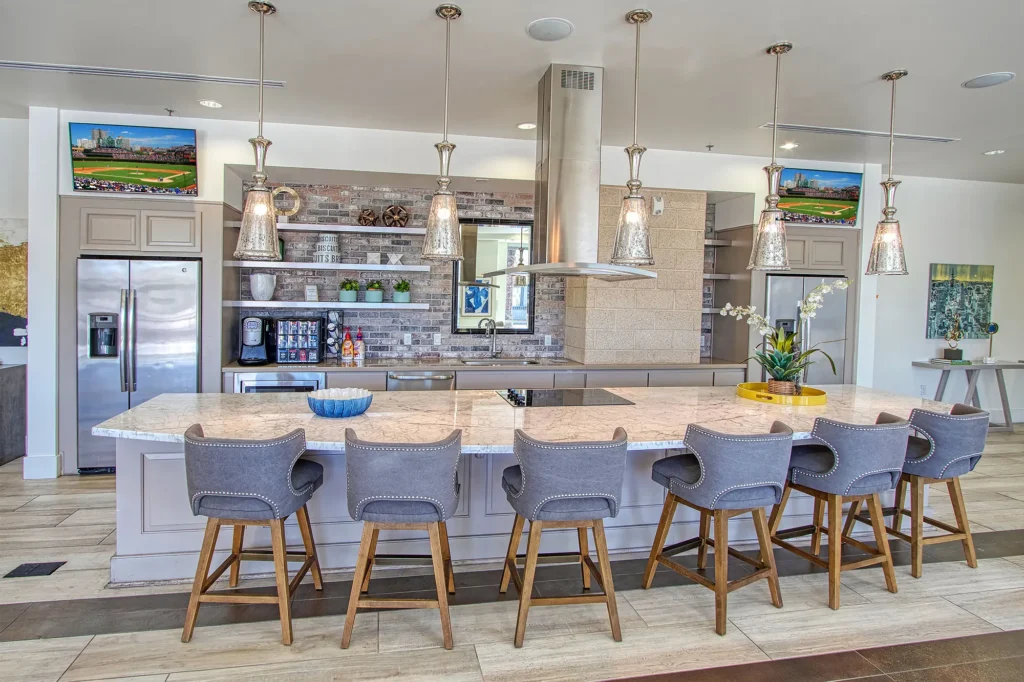 Modern clubhouse interior with a large kitchen island, barstool seating, stainless steel appliances, and lounge chairs facing kitchen space.