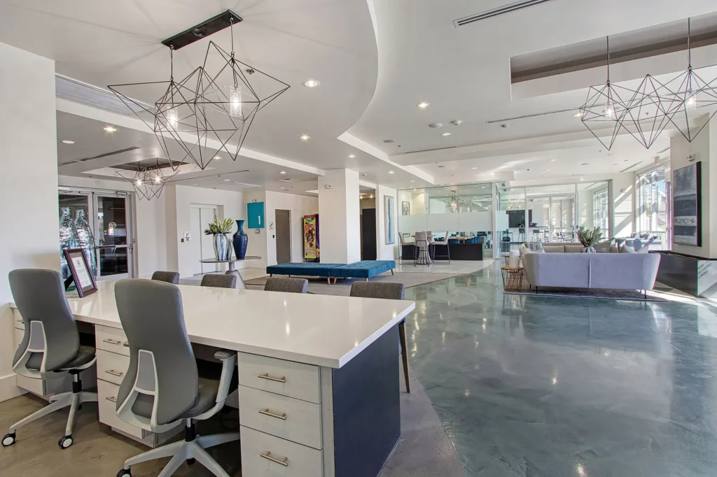 Modern office space with white desks, gray chairs, geometric light fixtures, a pool table, lounge area with sofas, polished floors, large windows, and contemporary decor. The environment is bright and spacious.