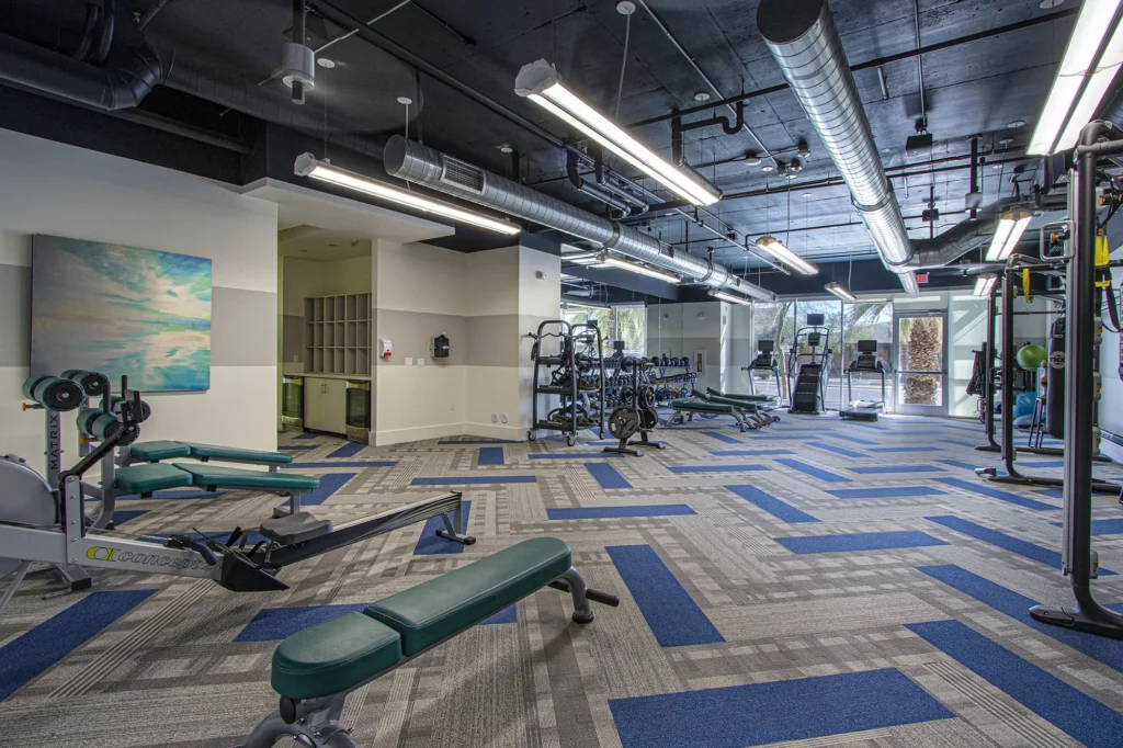 Carpeted fitness center with industrial style lighting and accents, with various strength and cardio training equipment.