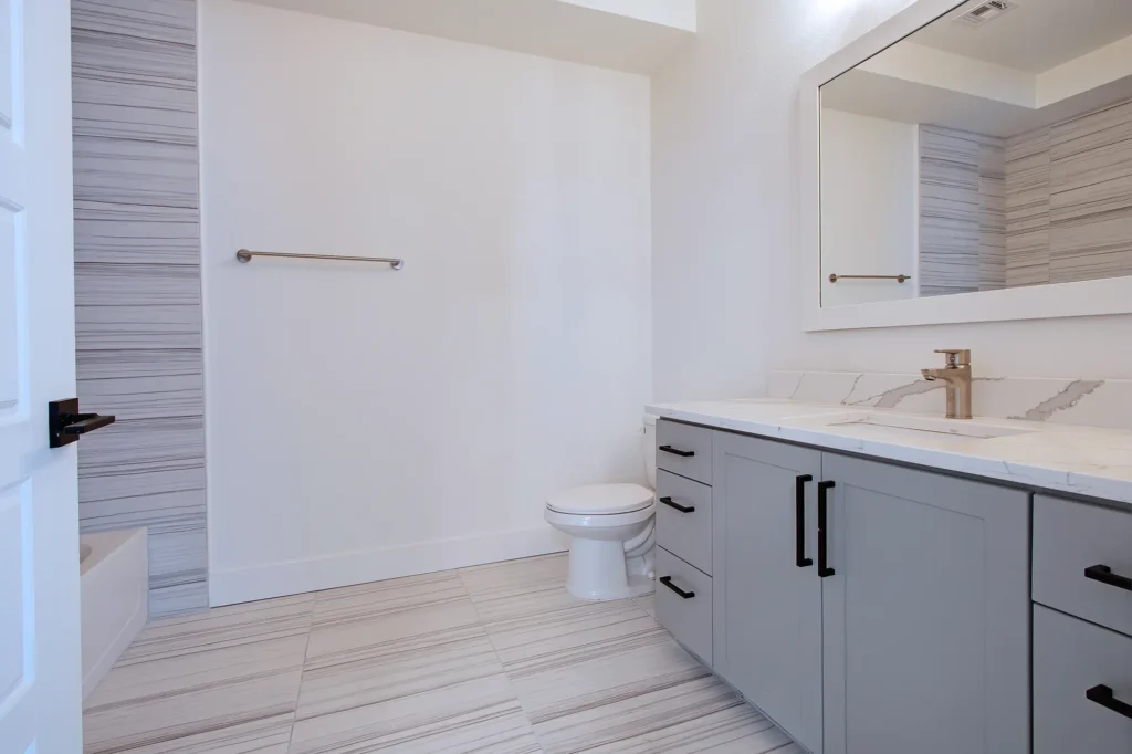 Modern bathroom featuring a gray vanity with a marble countertop, toilet, and elegant tile flooring. Natural light brightens the space.