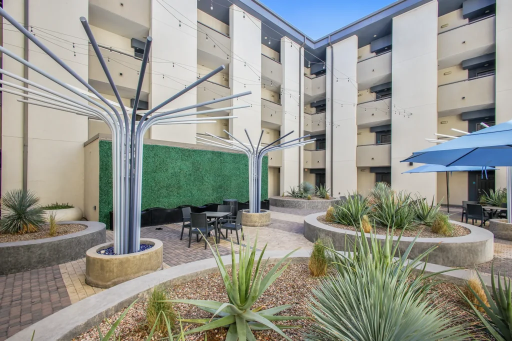Courtyard with desert landscaping, agave plants, outdoor seating under blue umbrellas, and modern metal sculptures.