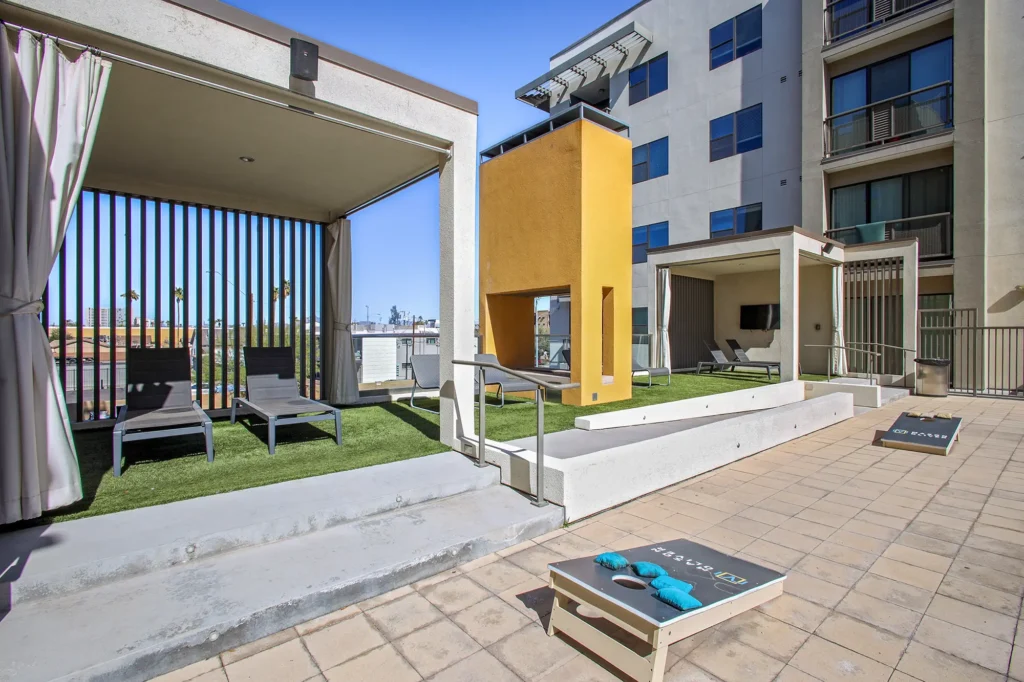 Outdoor lounge area with shaded cabanas, lounge chairs, a yellow outdoor fireplace structure, and cornhole game boards on the patio.