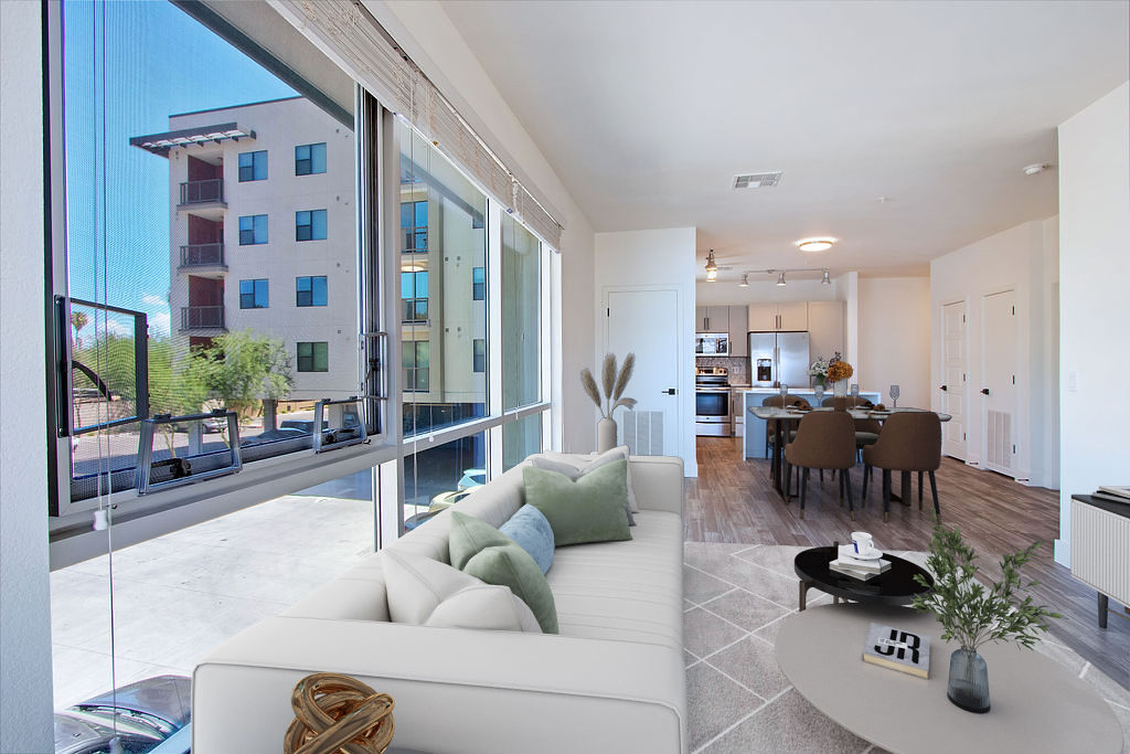A modern, bright apartment living room with a white sofa and green pillows, large windows, a dining area, and an open kitchen with stainless steel appliances. An apartment building is visible outside the window.