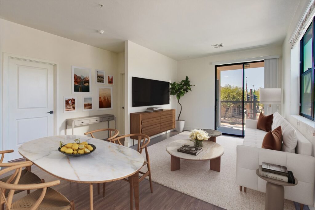 A modern living room with wood floors, a white sofa, dining area and seating, and a wooden sideboard. Large windows and a glass door let in natural light, and there’s a potted plant in the corner.