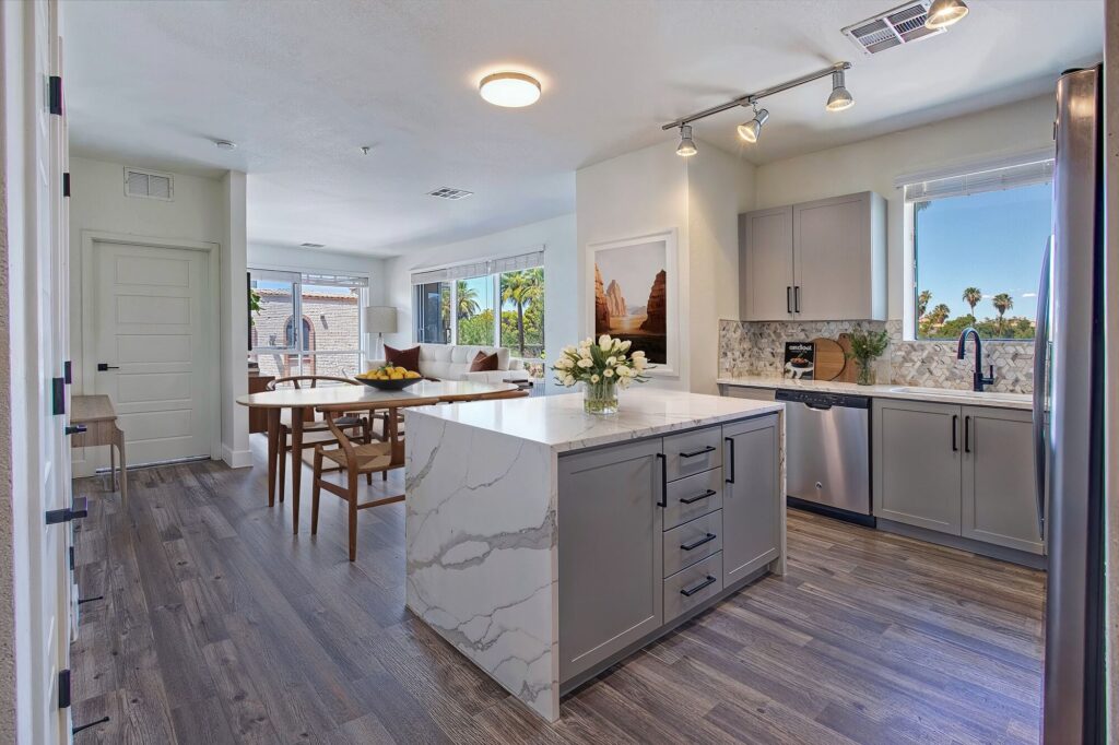 Modern kitchen with gray cabinets, marble island, stainless steel appliances, wood flooring, and dining area in the background. Large windows provide natural light and views of palm trees outside.