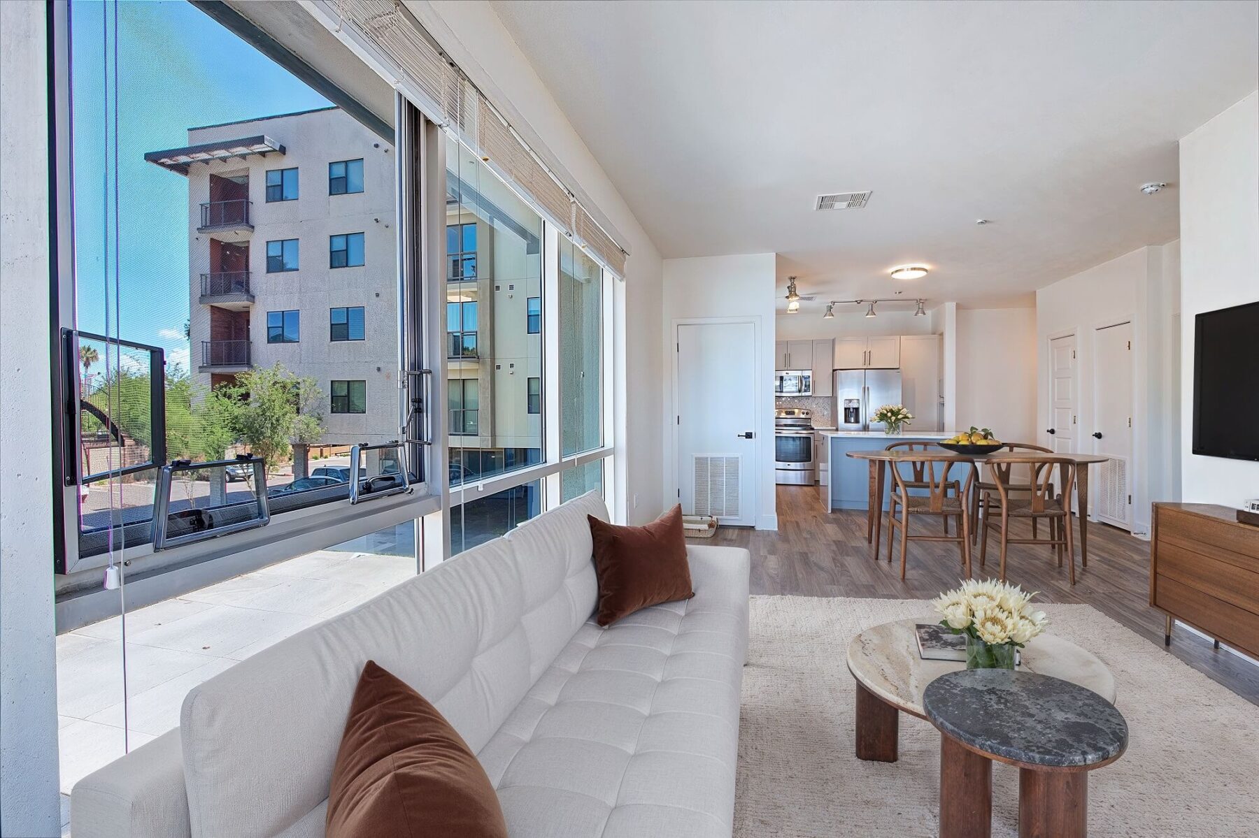 A modern, bright apartment living room with a white sofa and brown pillows, large windows, a dining area, and an open kitchen with stainless steel appliances. An apartment building is visible outside the window.