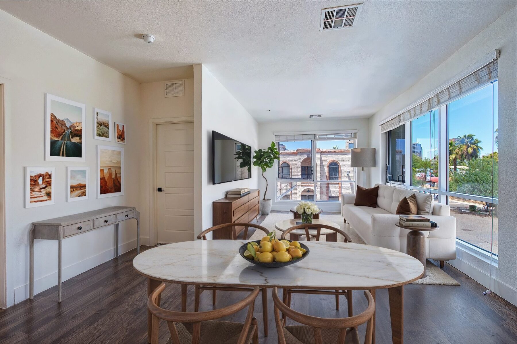 Modern apartment dining and living area with a marble table set for four, a cozy sofa, wall art, large windows with city and palm tree views, and bright natural light filling the space.