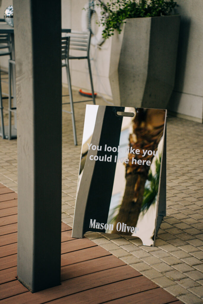 A mirrored sign on a tiled patio reflects palm trees and nearby furniture. The sign reads, You look like you could live here with Mason Oliver written at the bottom.