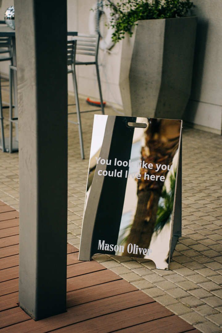 A mirrored sign on a tiled patio reflects palm trees and nearby furniture. The sign reads, You look like you could live here with Mason Oliver written at the bottom.