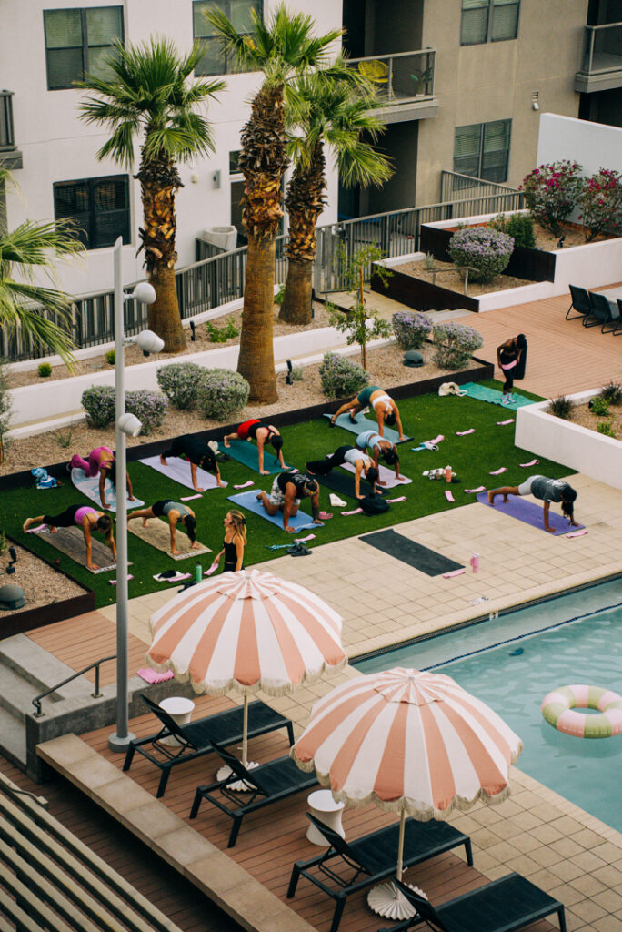 A group of people practice yoga on mats on a grassy area near a swimming pool, surrounded by palm trees and lounge chairs with striped umbrellas in an outdoor courtyard.
