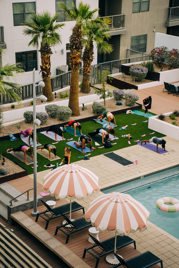 A group of people practice yoga on mats on a grassy area near a swimming pool, surrounded by palm trees and lounge chairs with striped umbrellas in an outdoor courtyard.