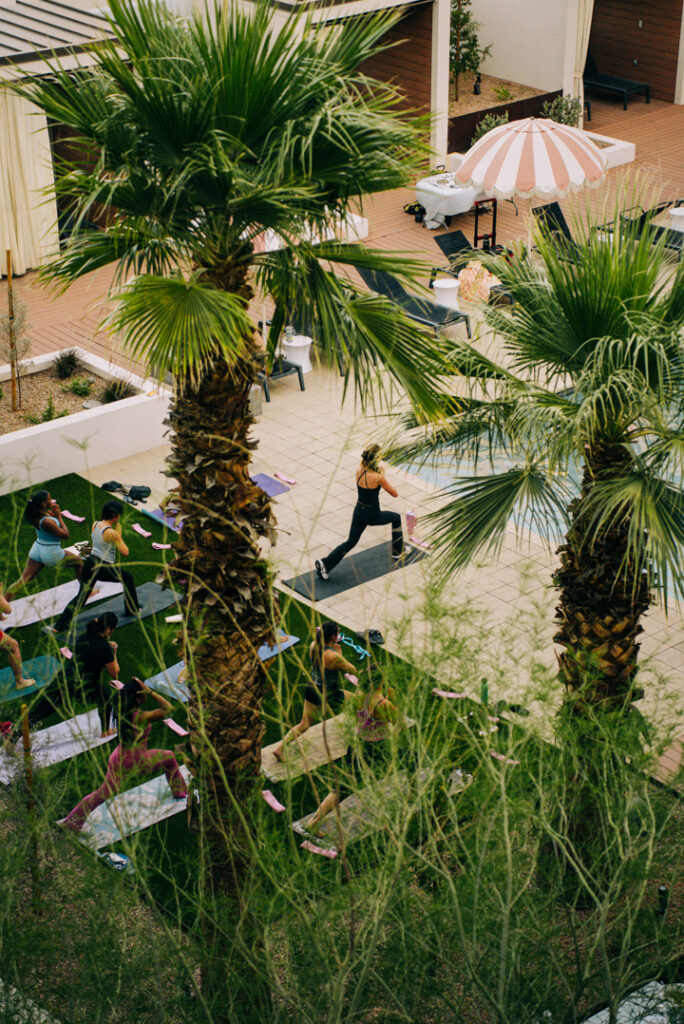 A group of people practice yoga outdoors on mats in a grassy area, surrounded by palm trees, with an instructor leading near a poolside under a striped umbrella.