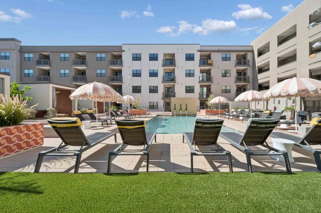 Modern apartment complex pool area with lounge chairs, pink-striped umbrellas, green artificial grass, and a multi-story building in the background under a blue sky with scattered clouds.