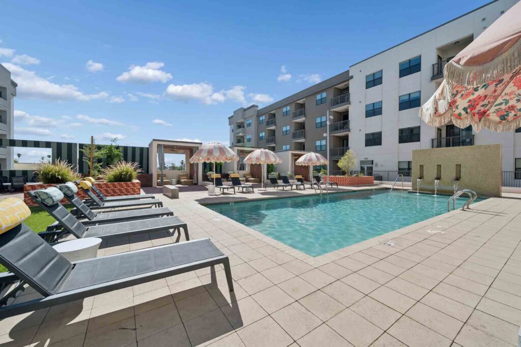 Outdoor apartment pool area with lounge chairs, umbrellas, cabanas, and potted plants. Modern apartment buildings surround the pool under a bright blue sky with scattered clouds.