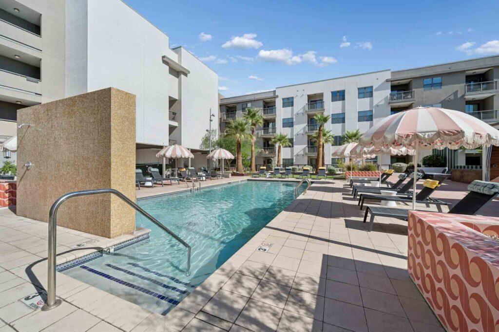 Outdoor pool with striped umbrellas, lounge chairs, palm trees, and modern apartment buildings in the background under a blue sky with scattered clouds.