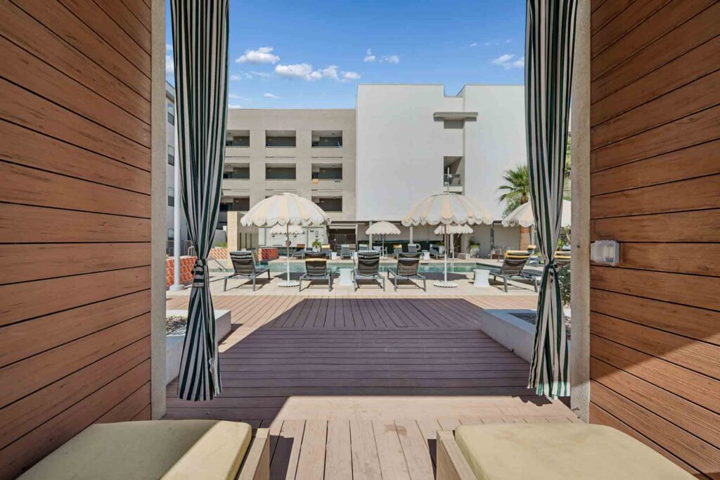 View from inside a cabana looking out to a pool area with striped umbrellas, lounge chairs, and a modern white building in the background under a blue sky.