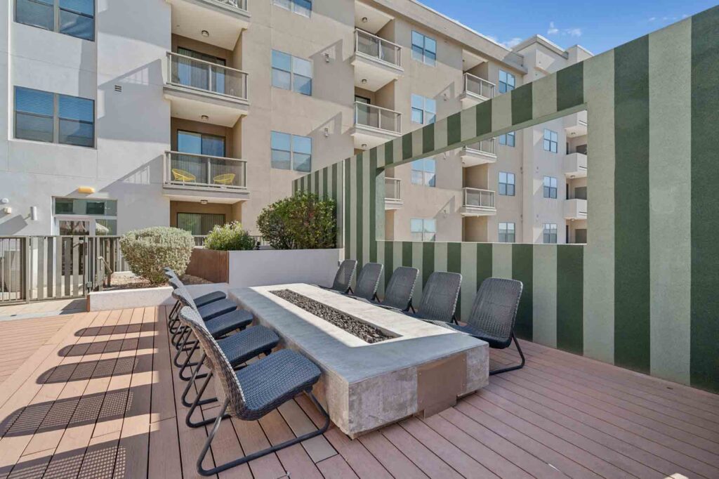 Outdoor patio with a long fire pit table surrounded by black chairs, wooden deck flooring, and vertical green-striped privacy wall, with modern apartment building in the background.