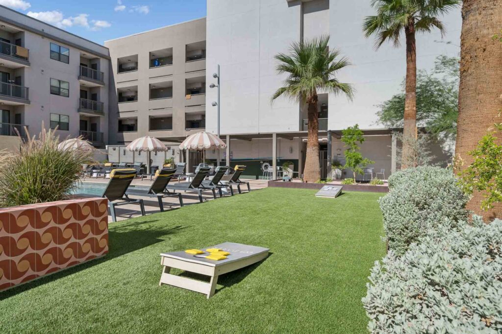 A grassy courtyard with cornhole boards, lounge chairs, palm trees, umbrellas, and surrounding apartment buildings on a sunny day.