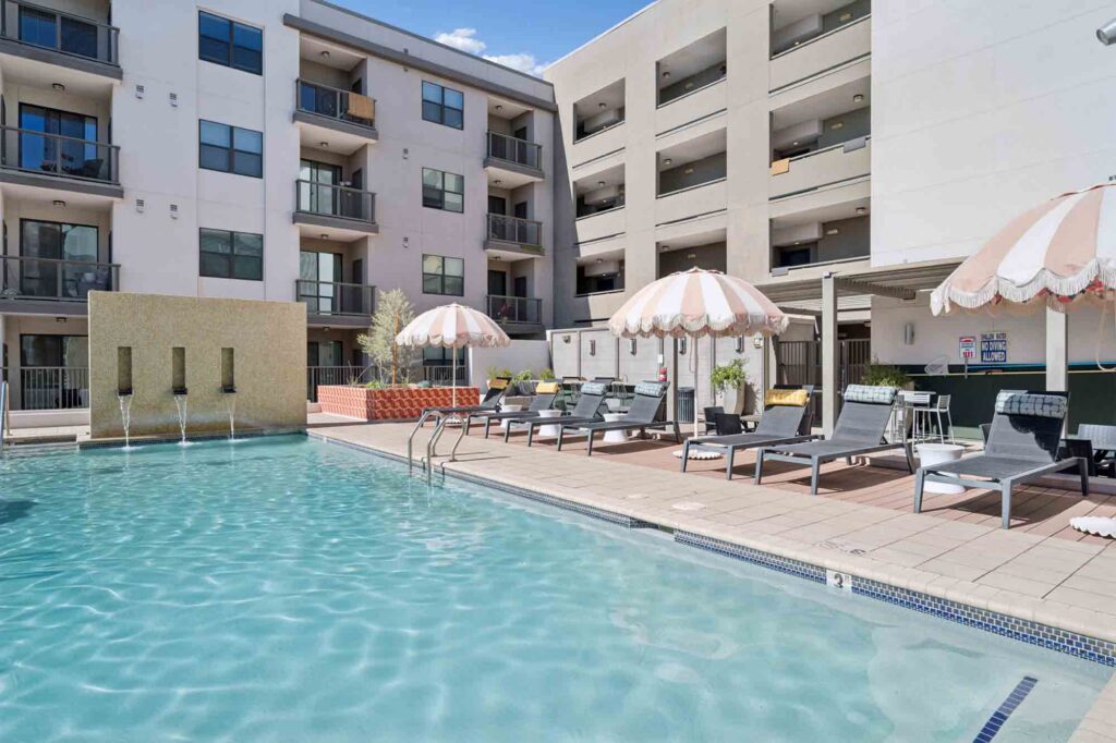 Modern apartment complex pool area with lounge chairs, striped umbrellas, and fountains. Multi-story buildings with balconies surround the pool, and the sky is clear and sunny.