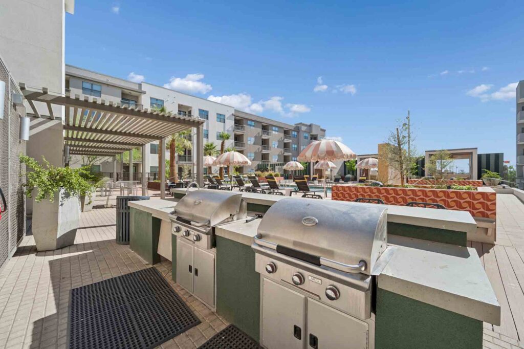 Outdoor lounge with two stainless steel grills, shaded seating under a pergola, lounge chairs with umbrellas, and modern apartment buildings in the background on a sunny day.