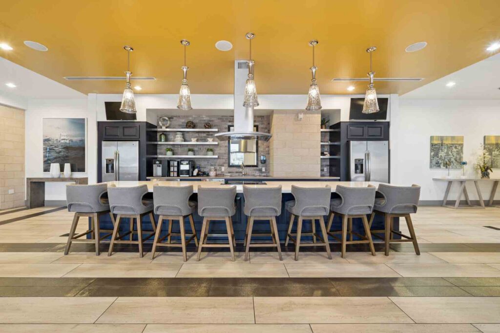 Modern kitchen with a long island, eight gray chairs, pendant lights, open shelving, dark cabinets, and a yellow ceiling accent. The space is bright with contemporary decor and clean lines.