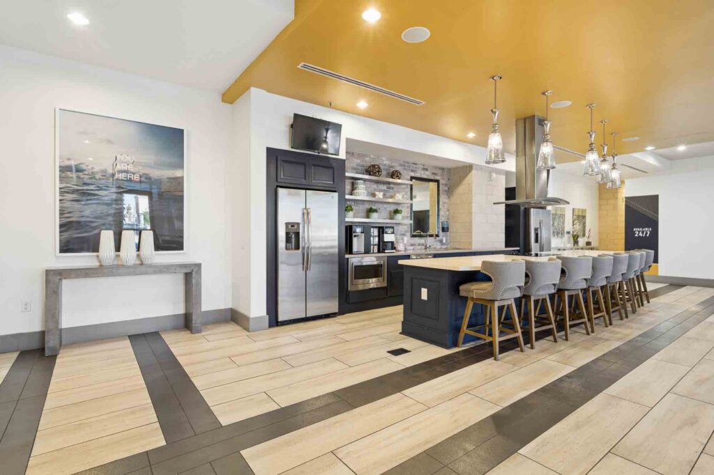 Modern communal kitchen with high chairs at a long island, stainless steel appliances, hanging lights, and yellow ceiling accents. Artwork and decor are visible on the left wall.