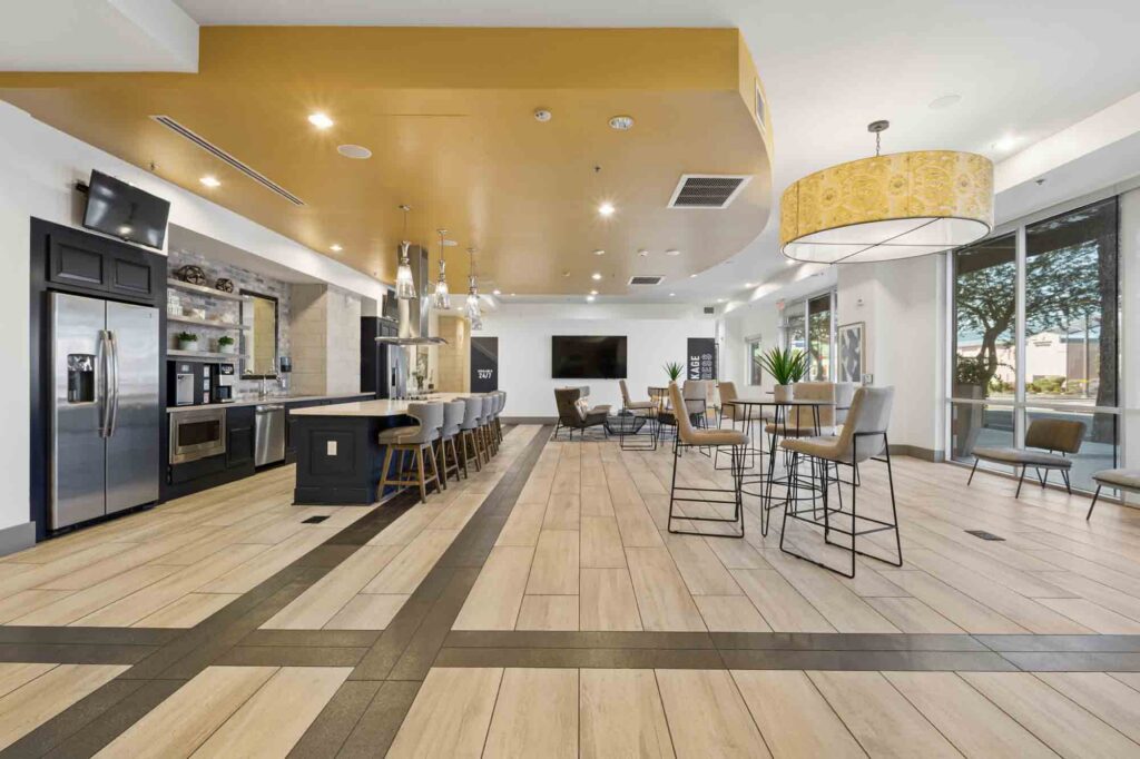 Modern communal kitchen and lounge area with high ceilings, island seating, hardwood floors, barstools, tables, large windows, and neutral decor. Stainless steel appliances and a yellow ceiling accent are visible.