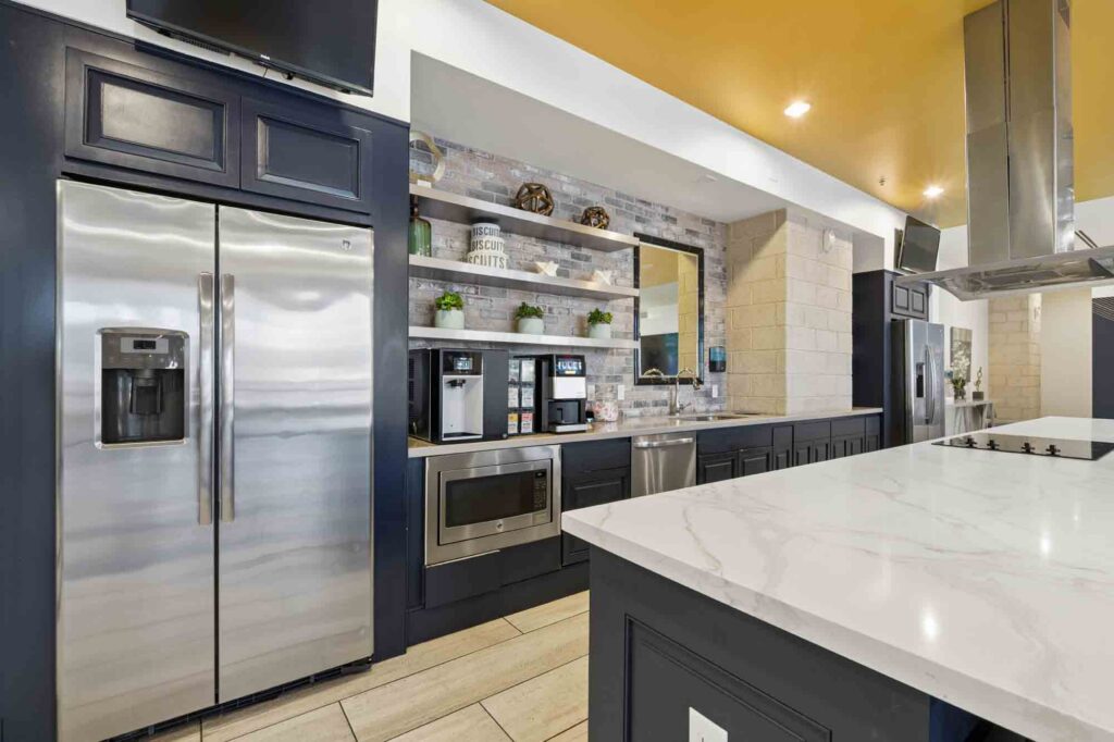 Modern kitchen with stainless steel fridge, microwave, coffee machines, open shelves with decor and plants, dark cabinets, and a large marble island countertop. Warm lighting and a yellow ceiling add brightness.