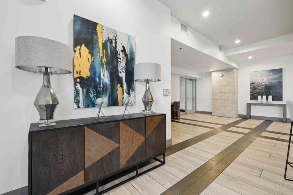 Modern hallway with wood and tile flooring, geometric sideboard, two silver lamps, abstract artwork, and a painting in the background near a doorway, all lit by recessed ceiling lights.