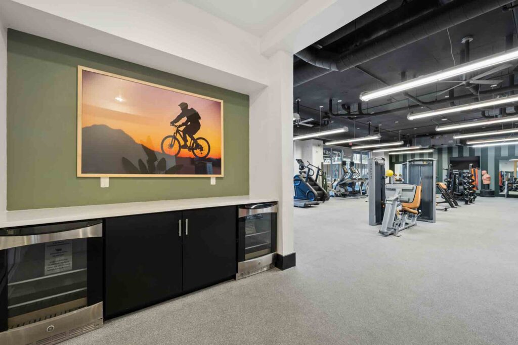 Modern gym with various exercise equipment, a green accent wall, and a framed picture of a cyclist at sunset above a cabinet with built-in mini fridges. Bright lighting and a clean, spacious layout.
