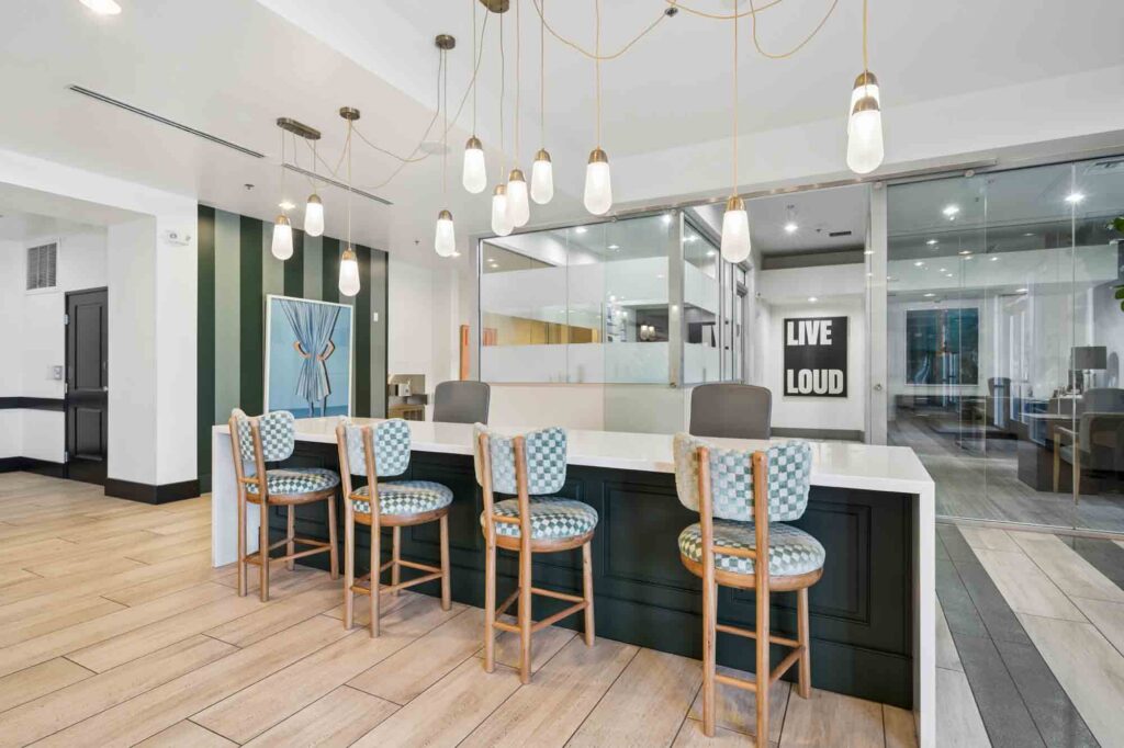 Modern communal workspace with four patterned chairs at a counter, hanging pendant lights above, glass-walled office in the background, and a Live Loud sign on the far wall. Light wood floor and bright decor.