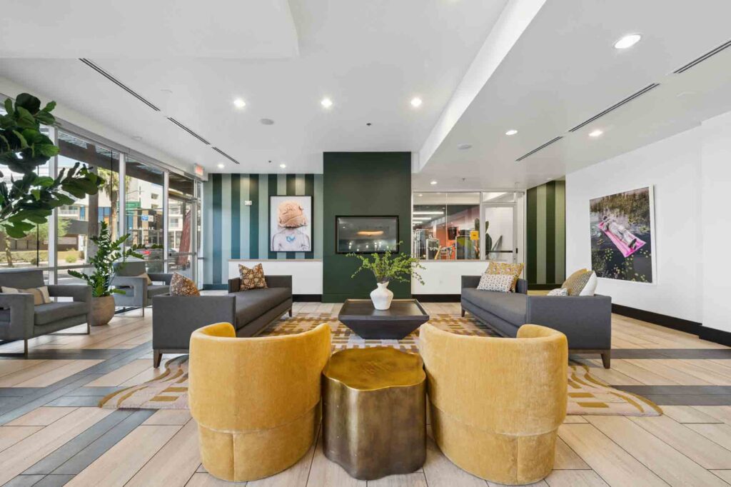 Modern lobby with yellow chairs, gray sofas, a central gold coffee table, potted plants, large windows, and wall art. A fireplace is built into a green accent wall. Bright, open, and contemporary design.