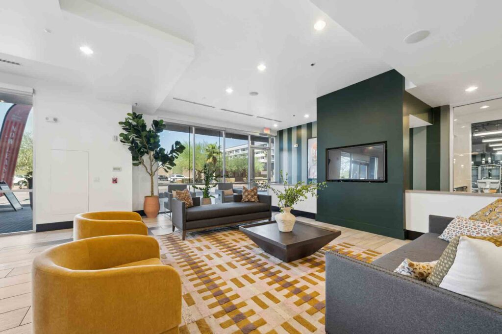Modern lobby with gold chairs, gray sofas, a patterned rug, large windows, and green accent wall with a TV. Potted plants and natural light create a welcoming atmosphere.