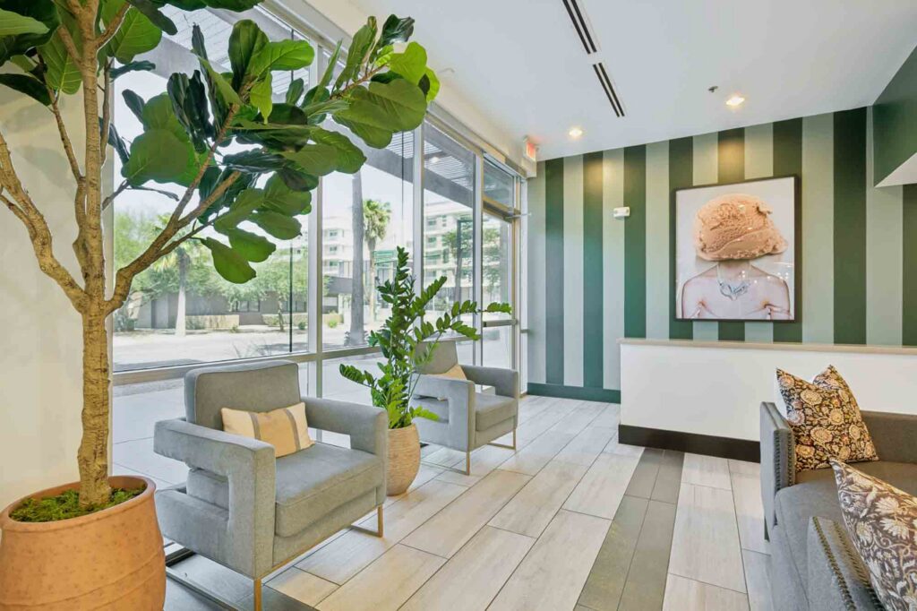 Modern waiting area with potted plants, gray chairs with cushions, and a striped accent wall. Large windows let in natural light; a framed portrait hangs on the wall above the seating area.