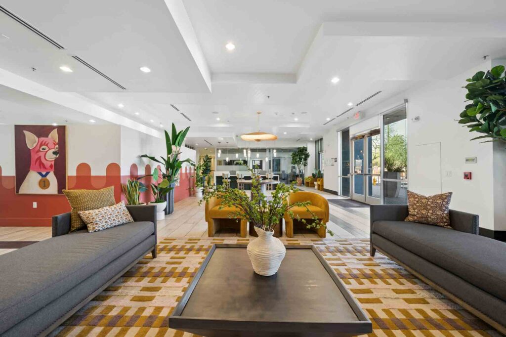 Modern hotel lobby with gray sofas, yellow chairs, a coffee table with a white vase, decorative pillows, plants, large windows, and colorful wall art featuring a cartoon dog. Natural light fills the space.