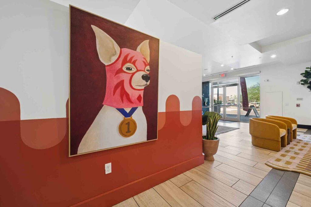 A large painting of a dog wearing a pink mask and a gold medal hangs on a color-blocked wall in a modern lobby with tan chairs, plants, and natural light from glass doors.