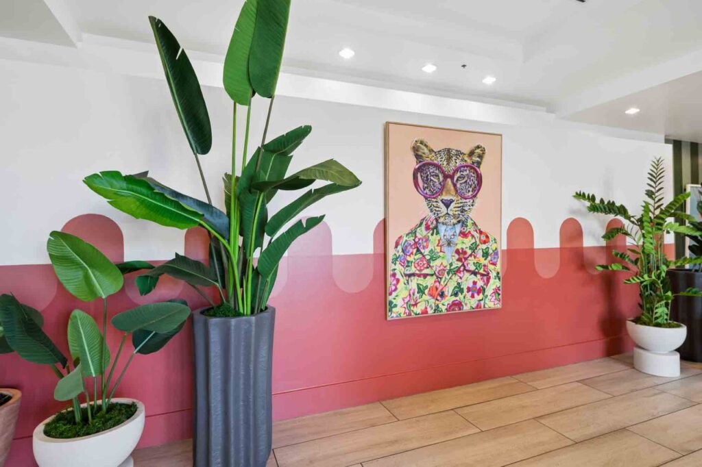 Large green potted plants sit on a wood floor near a white and red wall. A colorful framed painting of a cat in floral clothes and round glasses hangs on the wall. Ceiling lights are visible above.