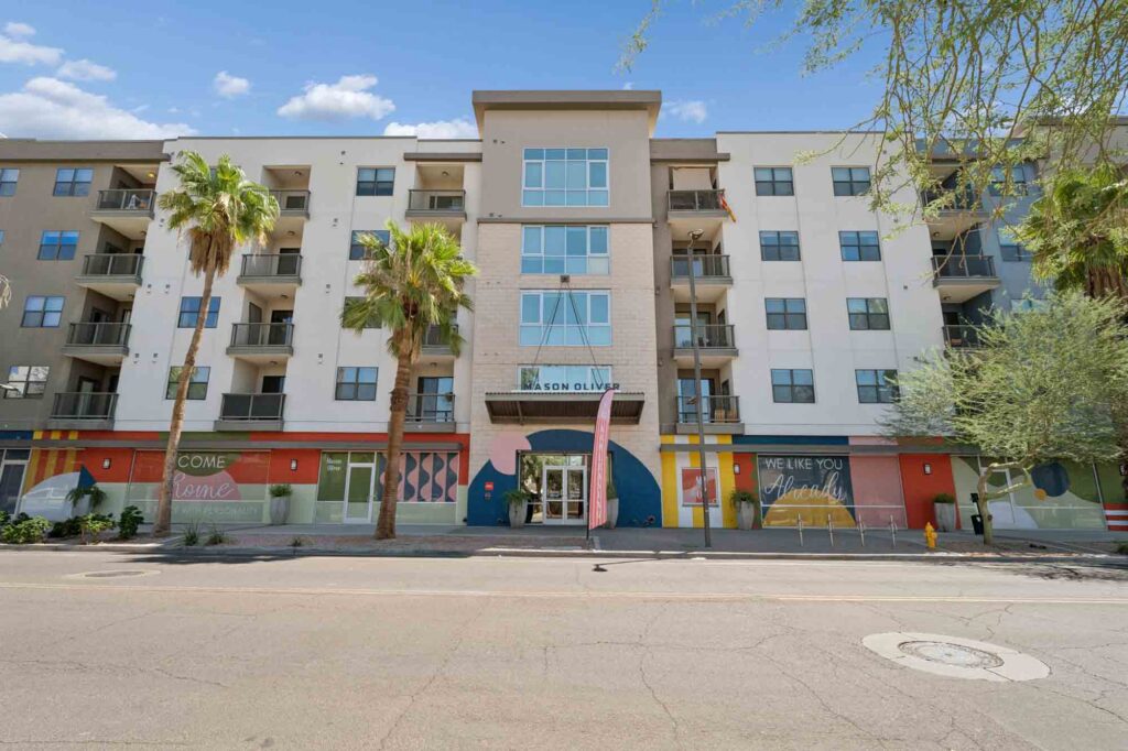 Modern four-story apartment building with balconies, palm trees in front, and colorful murals at street level. Blue sky and some clouds overhead.