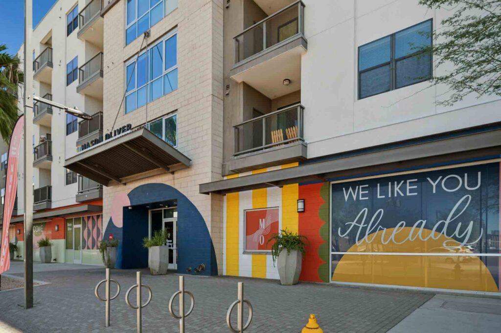 Modern apartment building exterior with large windows, plants in tall pots, and colorful mural on the wall. The mural reads WE LIKE YOU Already in big letters. Bike racks and a sidewalk are in front.