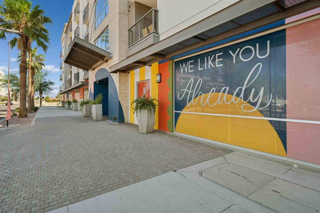 A modern building with colorful wall art that says WE LIKE YOU Already, large potted plants, palm trees, and a paved sidewalk under a blue sky.