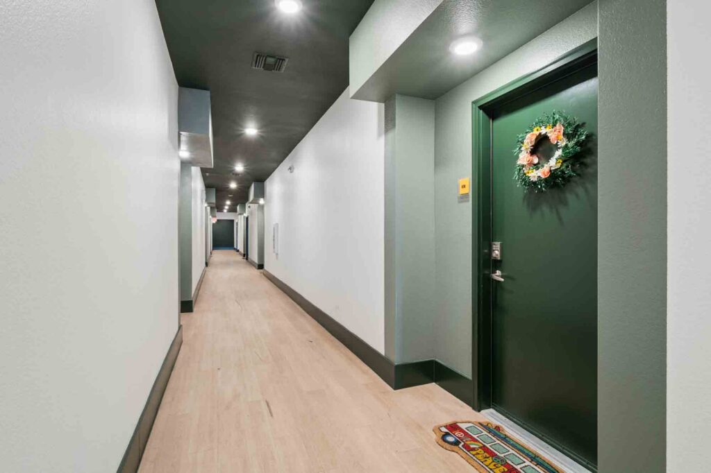 A brightly lit apartment hallway with light wood floors. One dark green door is decorated with a wreath and a colorful welcome mat. The walls are pale gray and the ceiling has several recessed lights.