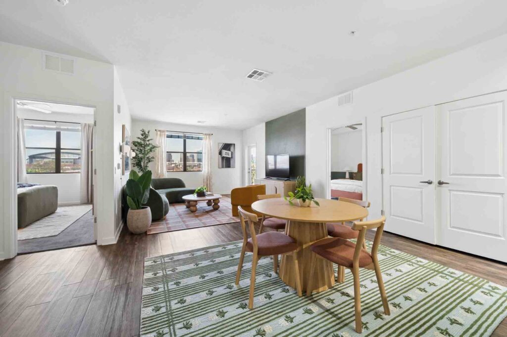 Modern apartment interior with a round dining table, four chairs, living area with a green sofa, TV, and large windows. Two bedrooms and white double doors are visible. Green and white rug on the floor.