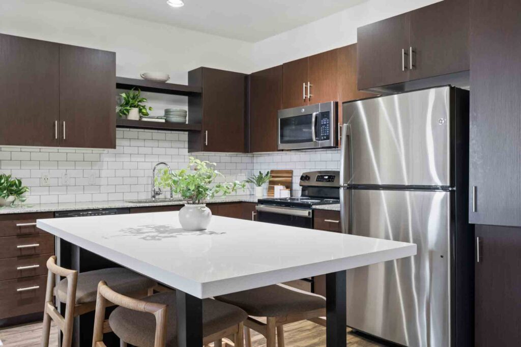 Modern kitchen with dark wood cabinets, white subway tile backsplash, stainless steel appliances, a white island with four chairs, and plants as decor.