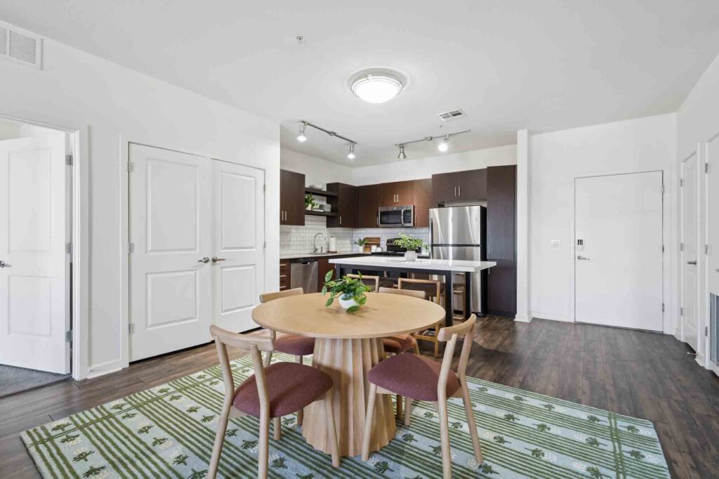 Modern kitchen and dining area with a round wooden table, four chairs, a green rug, stainless steel appliances, and dark cabinets. White walls and doors provide a bright, clean atmosphere.