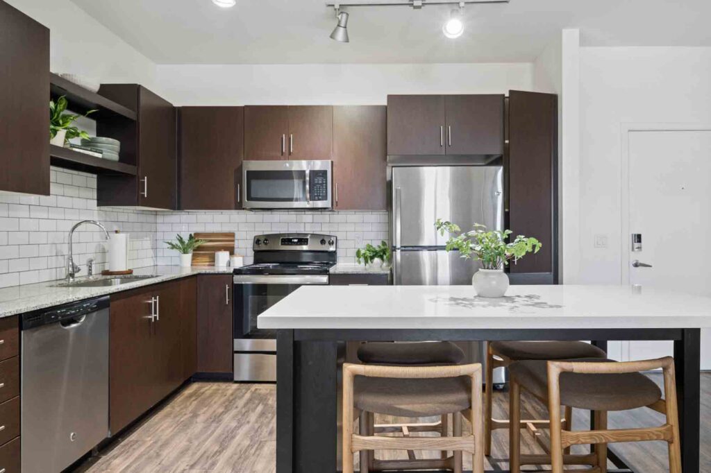 Modern kitchen with dark cabinets, stainless steel appliances, white tile backsplash, a large island with barstools, potted plants, and wood flooring. Bright, clean, and organized space.