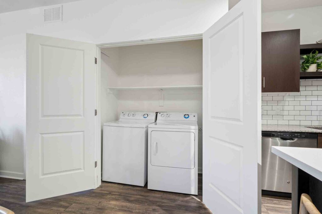 A laundry closet with white bi-fold doors, containing a white washing machine and dryer side by side, with a shelf above them. The closet is in a modern home with wood flooring and nearby kitchen appliances.