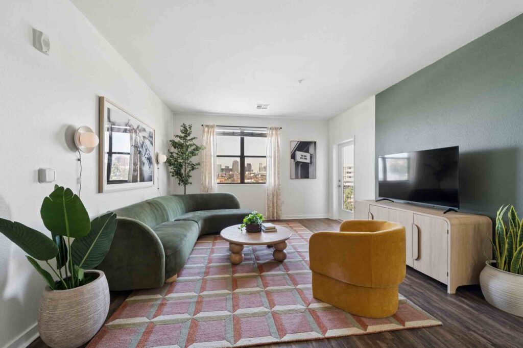 Modern living room with green sofa, yellow chair, geometric rug, potted plants, large wall art, and TV on light wood console; window lets in natural light.