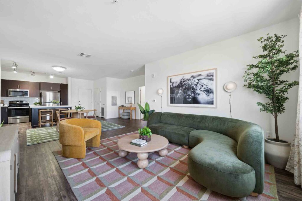 Modern living room with a green curved sofa, yellow chair, round coffee table, patterned rug, plant, wall art, and open kitchen in the background with dark cabinets and stainless steel appliances.