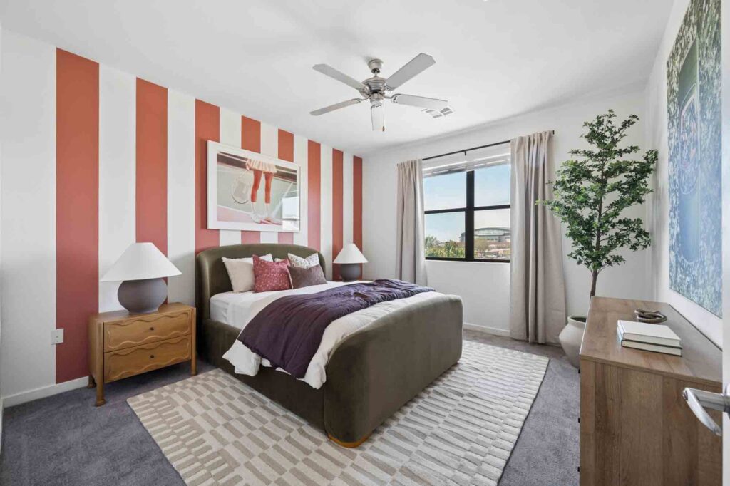 Modern bedroom with red and white striped accent wall, bed with purple accents, bedside tables with lamps, large window with beige curtains, potted plant, and dresser with books and decor.
