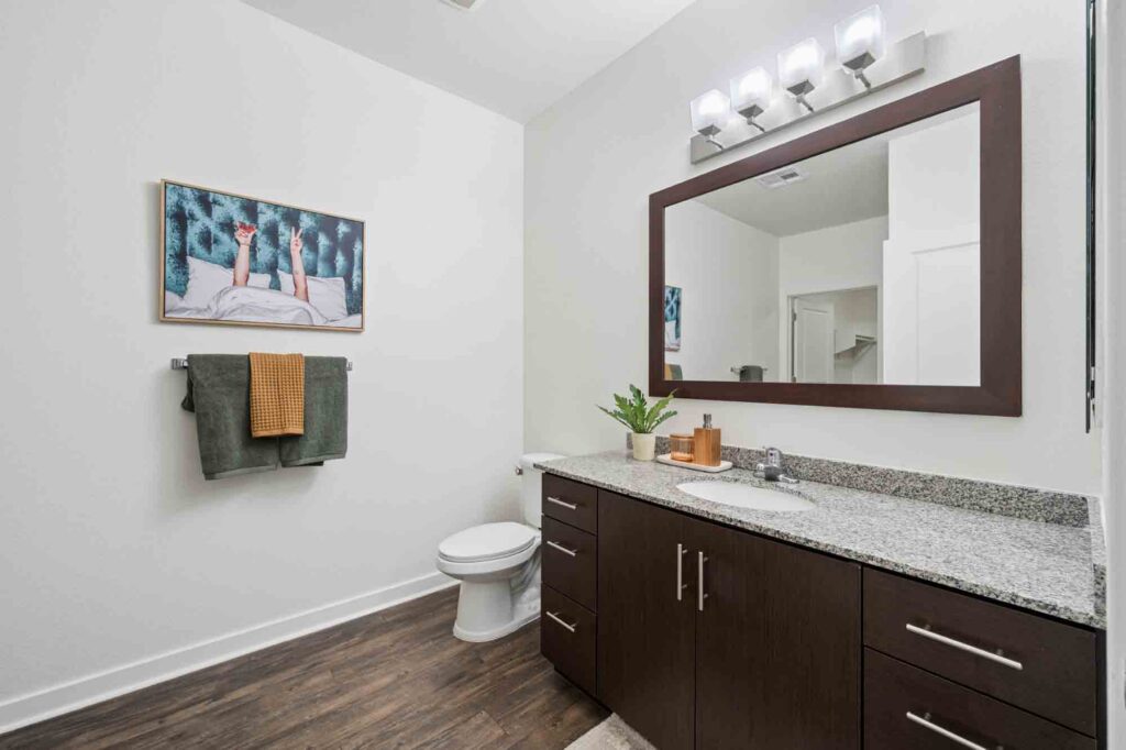Modern bathroom with dark wood cabinets, granite countertop, large mirror, white walls, hardwood floor, towel rack with green and brown towels, toilet, and framed wall art above the towels.